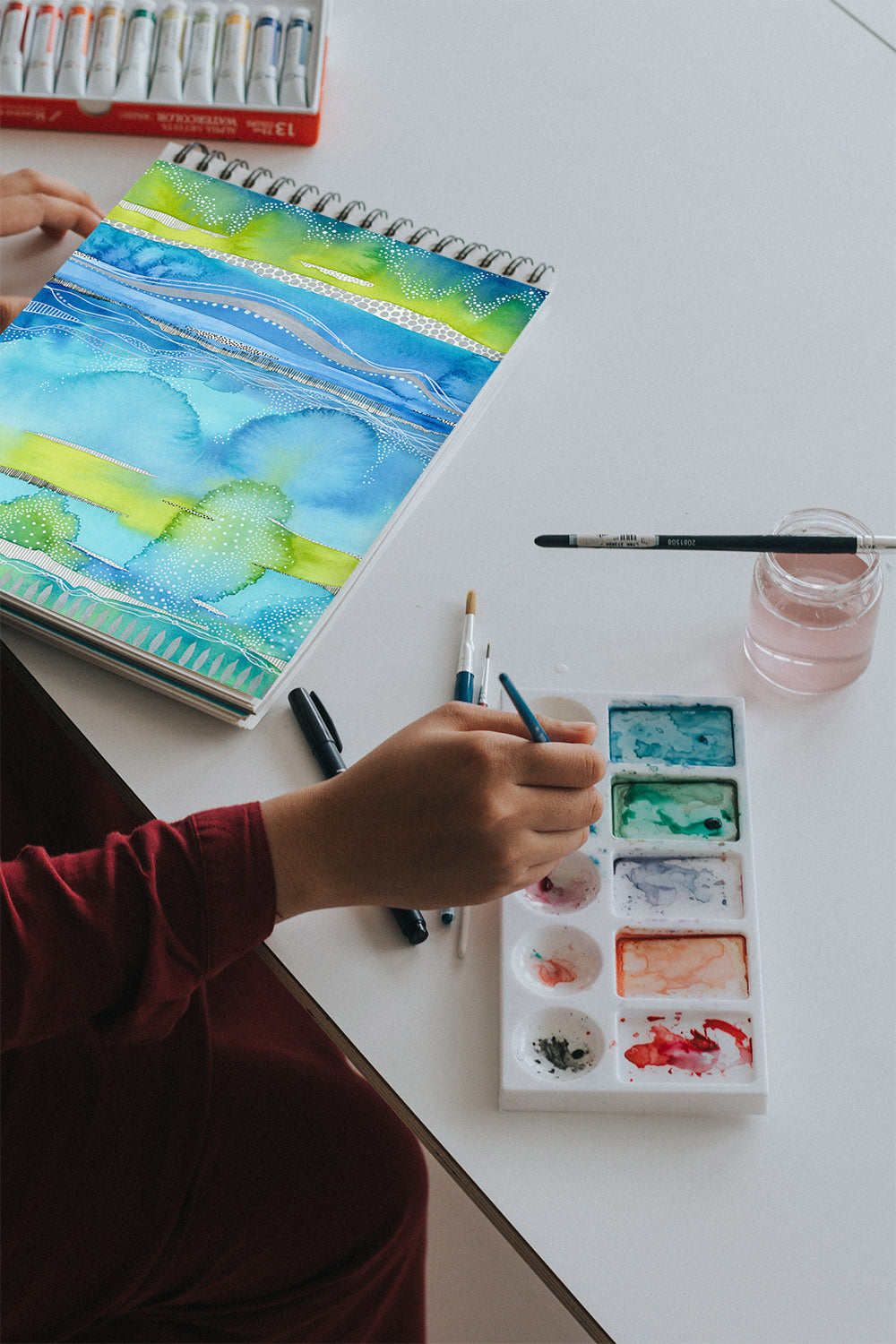 this is an image of a person sitting at a table painting an abstract landscape painting using blue, teal and lime green.  the person is dipping their paint brush into a pan of watercolor paints.  there is also a jar a water with a paint brush laying over it in front of the person.
