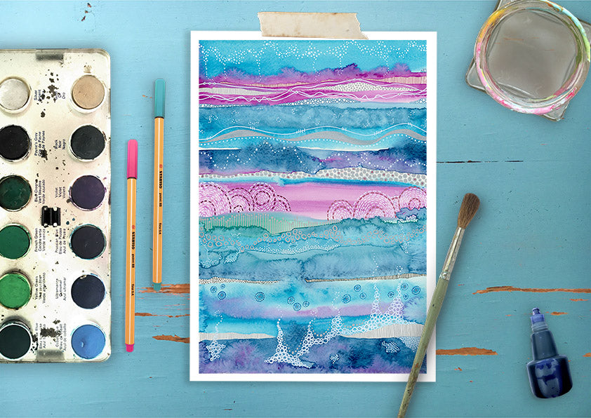 This image contains a watercolor painting by joanne grant art.  The painting is an abstract landscape in teal, pink and dark blue.  There are lines, circles and dots in white, pink and blue ink.  The painting is on a wood blue table with a watercolor palette, pens, blue ink bottle, a brush and a glass water jar surrounding it.