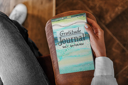 This is an image of a person sitting on a chair holding a gratitude journal on the arm of the chair.  The image is cropped so you only see a portion of the leg and arm of the person and a portion of the arm of the chair.  The gratitude journal is by joanne grant art.  It is volume 1.  The cover of the journal is a painting by joanne grant.  It is blue and teal with shades of green.