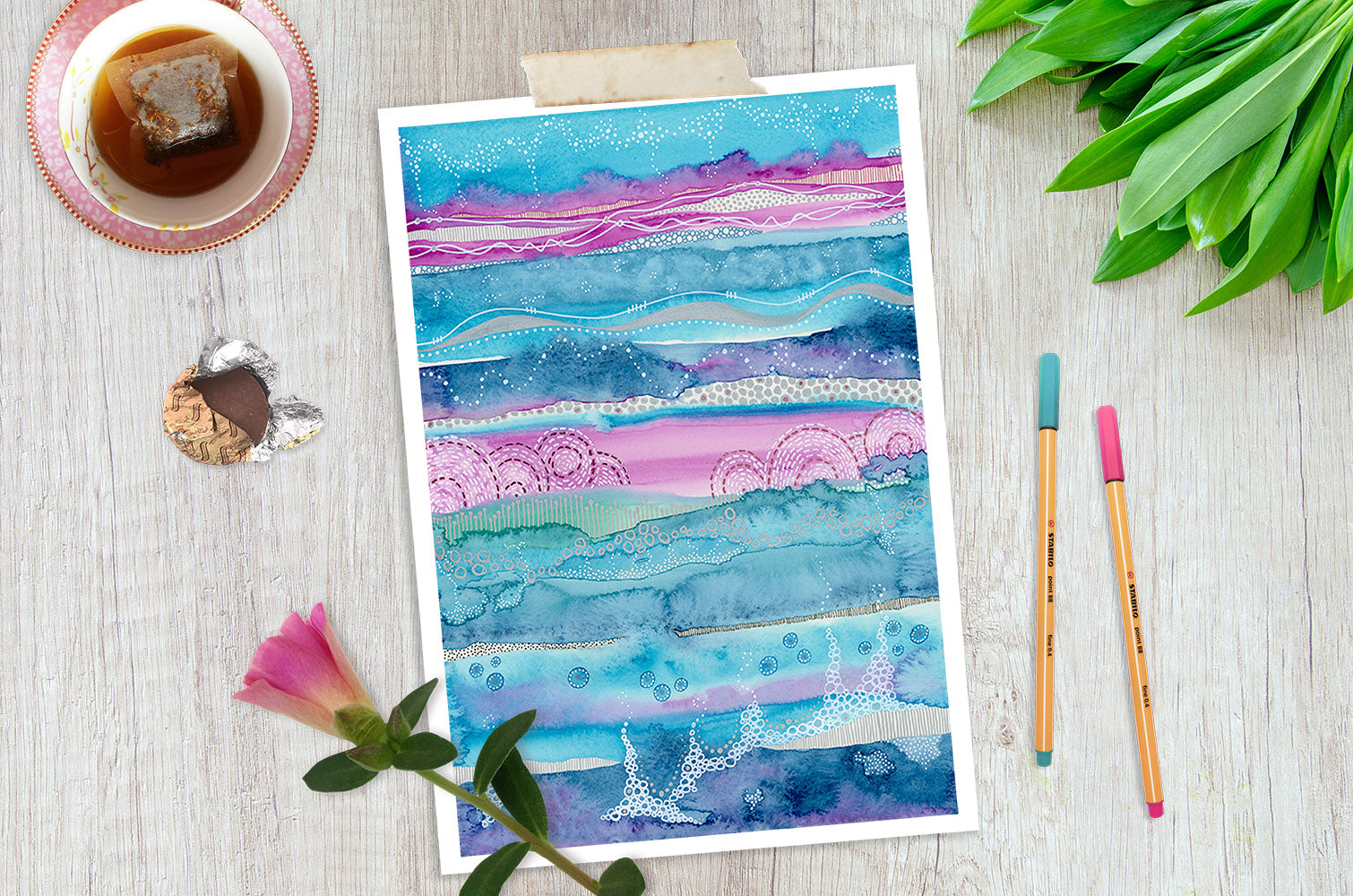 This image contains a watercolor abstract landscape painting in blue and pink by joanne grant art.  The painting is on a white wood table with a cup of tea, a chocolate, pink flower, teal and pink pens and a green plant surrounding it.
