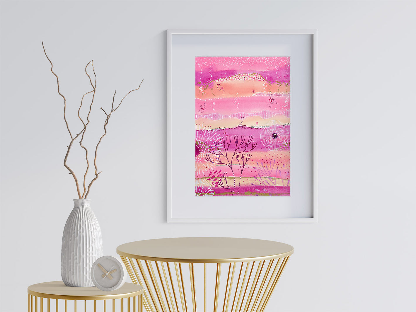 This image is an abstract painting of a coral reef done by joanne grant art.  The painting is shades of pink.  In white ink there are abstract under sea flora.  Many abstract coral reef plants are in the painting also.  The painting is framed in a white frame and it is hanging on a white wall.  There are two gold modern metal tables in the scene in front of the painting.  On the left table is a white textured vase with several dried branches extending from it.  There is also a clock on that table.   