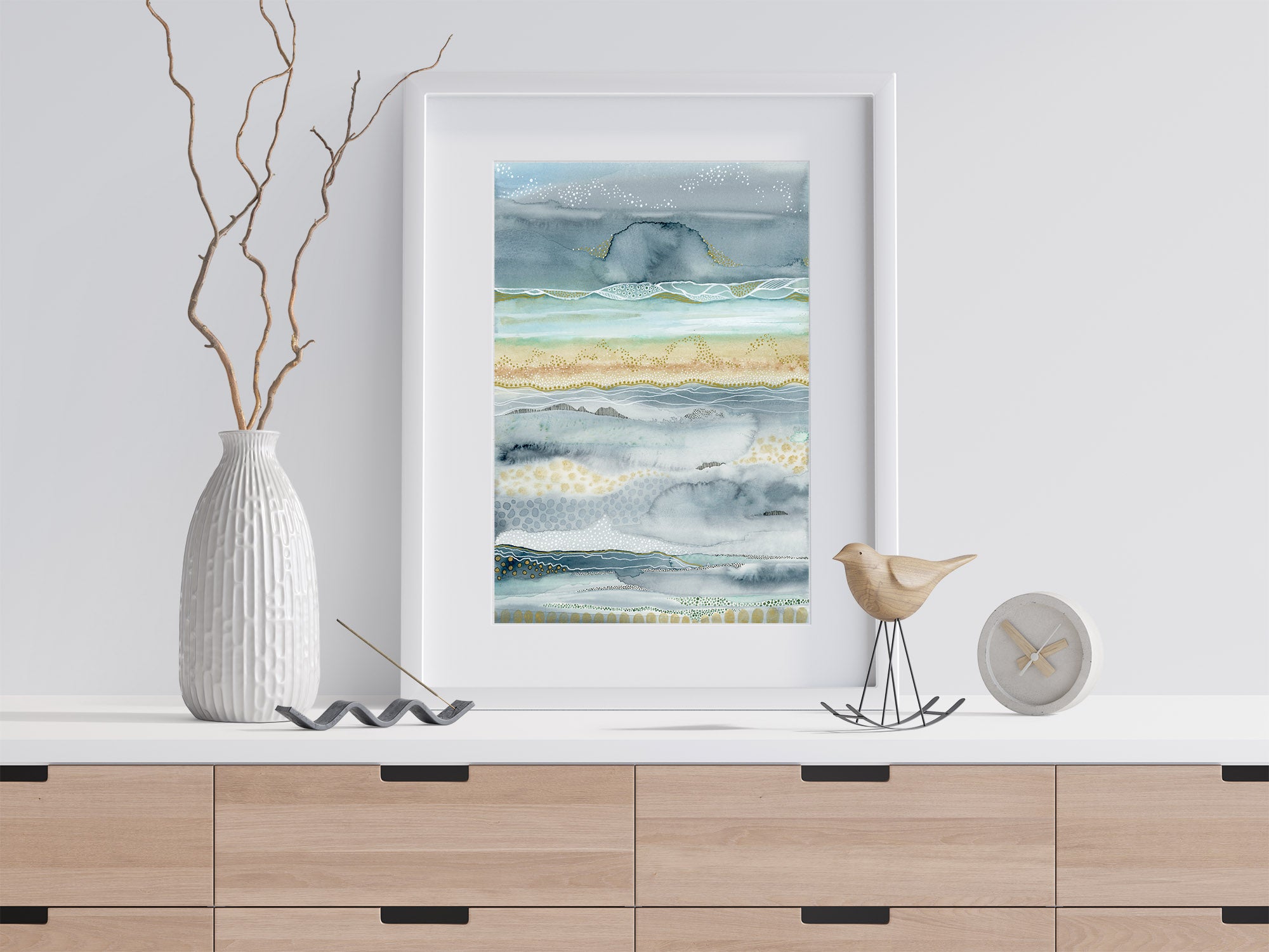  The is an image of a watercolor painting done by joanne grant art.  The painting is shades of green and teal and it is framed in a white frame.  The painting is sitting on a light wood table with a white top leaning against the wall.  To the left of the painting is a white textured vase with dried branches extending out of it.  To the right of the painting is a wood bird statue.  Next to that is a small clock.