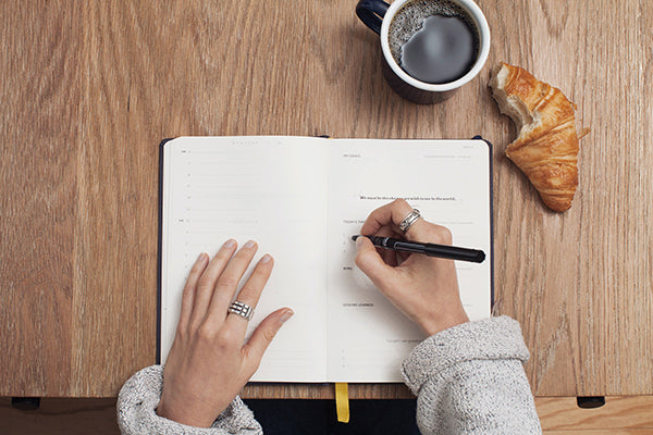 This is an image of a looking downward to a desk top.  A girl's hands are writing in a notebook which is on the desktop. There is a mug of coffee and a croissant with a bite taken out of it to the top right of the book.