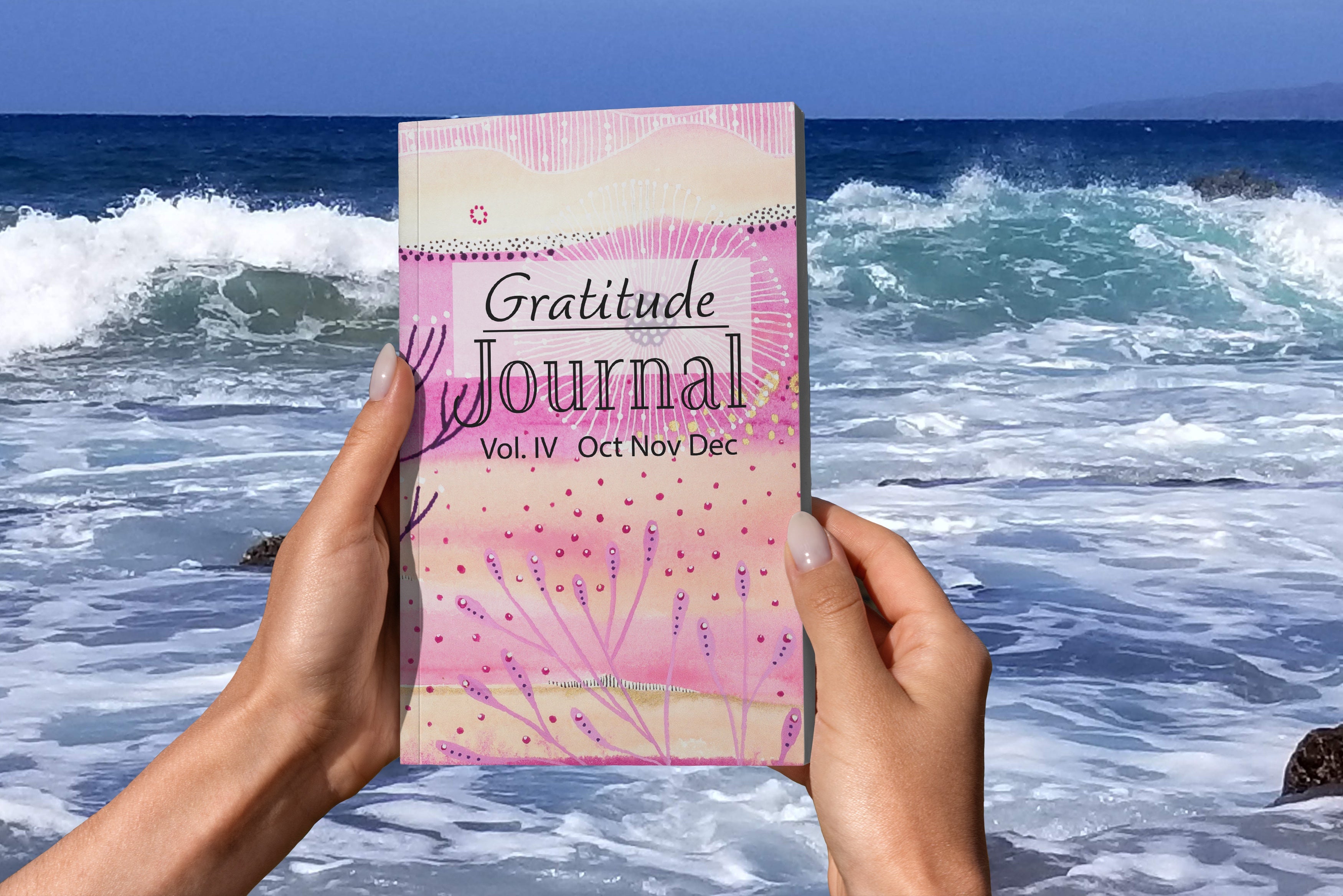hands holding gratitude journal written by joanne grant.  This gratitude journal is volume 4, October, November and December.  The hands are holding the gratitude journal in front of waves crashing.