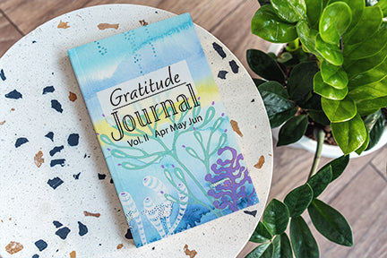 This is an image of a gratitude journal by joanne grant.  The journal's cover is an image of a watercolor painting by joanne grant.  The watercolor painting is of a whimsical coral reef painted in blue, green and purple.  The book is sitting on a white round stone table.  There is a large green plant in the image.
