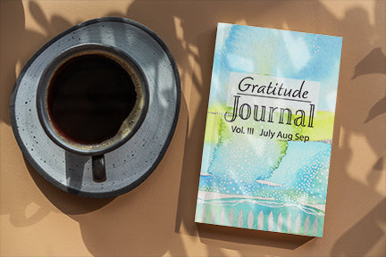 This image contains a gratitude journal by joanne grant art sitting on a light brown table with a cup of coffee to the left of the book.  The gratitude journal's cover has a watercolor abstract landscape painting by joanne grant on it.  The colors are blue, teal, and lime green.  This is vol 3 of the gratitude journal.