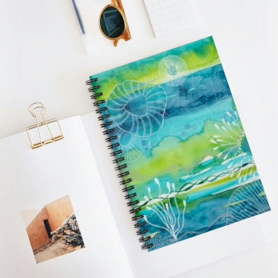 This is an image of a spiral note book laying on a white table.  The notebook features an abstract coral reef painting in teal, blue and lime green painted by joanne grant.  To the left and under the notebook is a white open book with a gold clip holding the pages open.  Above the books are a pair of tortoise shell sunglasses folded closed.